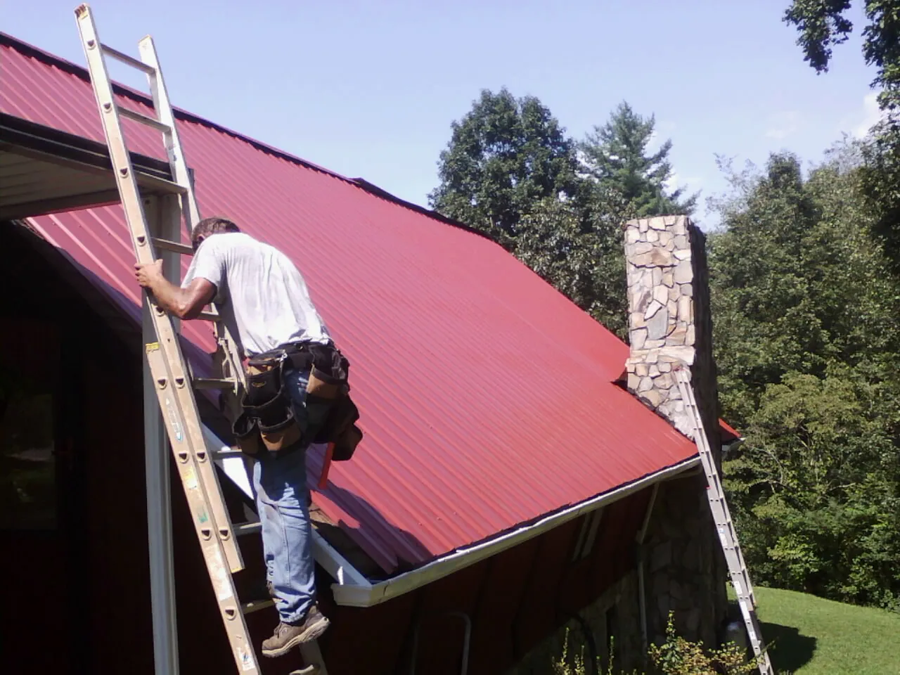 Roofer on a ladder beside a red metal roof and stone chimney — Soffit & Fascia Repair by Roofing Brothers in Le Grand